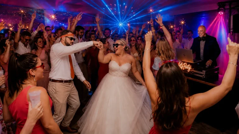 Bride and groom dancing on a packed wedding dancefloor with guests celebrating and a professional wedding DJ performing under elegant blue and pink lighting.